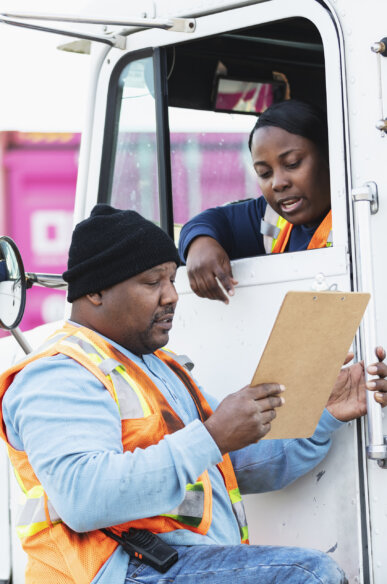 A truck driver woman talks to a worker wearing an orange vest while holding a clipboard on his hands
