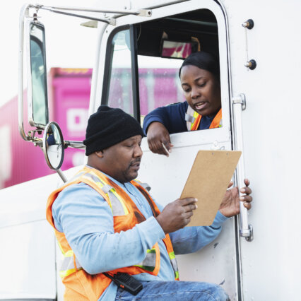 A truck driver woman talks to a worker wearing an orange vest while holding a clipboard on his hands