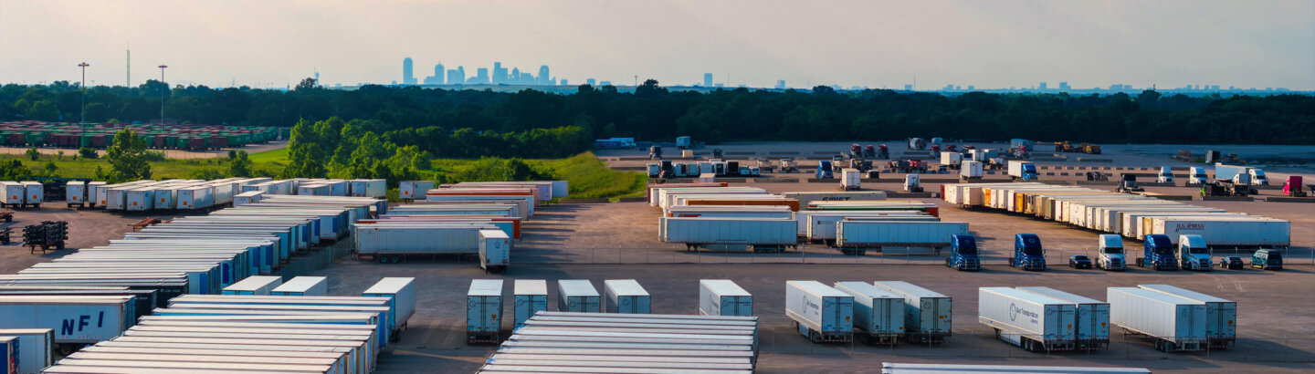 Rows of parked trailers and trucks in a lot with the city skyline in the distance